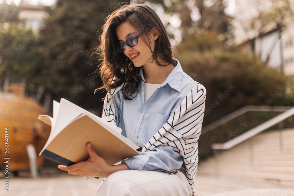 Fototapeta premium Attractive young caucasian lady leafing through book preparing for exam sitting near college. Brunette with wavy hair wears blue shirt. Outdoor education concept