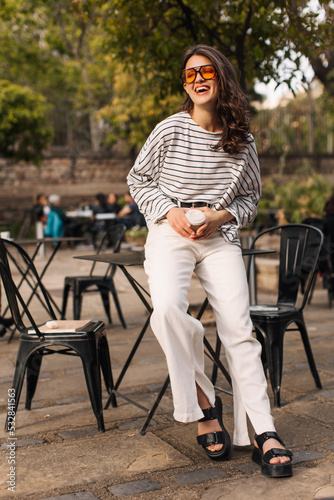 Full length happy young caucasian woman laughing relaxing in outdoor cafe spring. Brunette with wavy hair wears casual clothes. City life concept