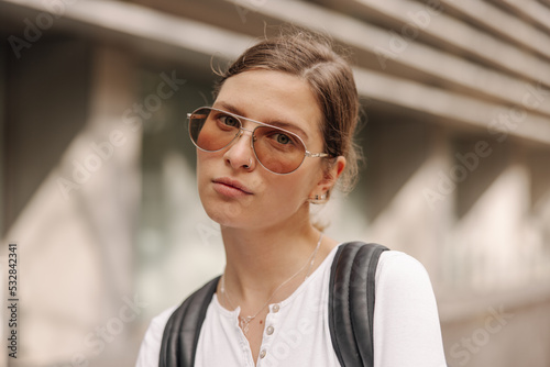 Close-up of young caucasian woman in sunglasses looking at camera on blurred background. Serious brown-haired wears white jacket and backpack her shoulders. City life concept