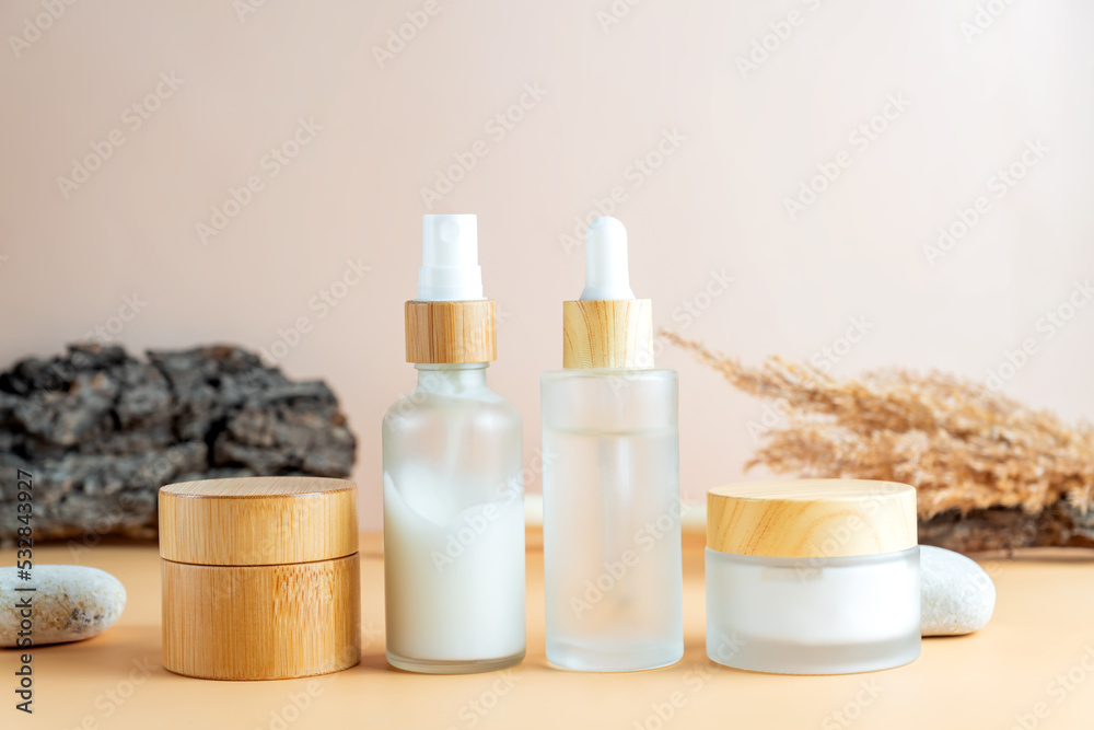 White frosted glass cosmetics bottles and jars with pamboo lids with tree bark, pampas grass, stones on beige background. Autumn fall beauty products set. White frosted glass cosmetics bottles and jars with pamboo lids with tree bark, pampas grass, stones on beige background. Autumn fall beauty products set.