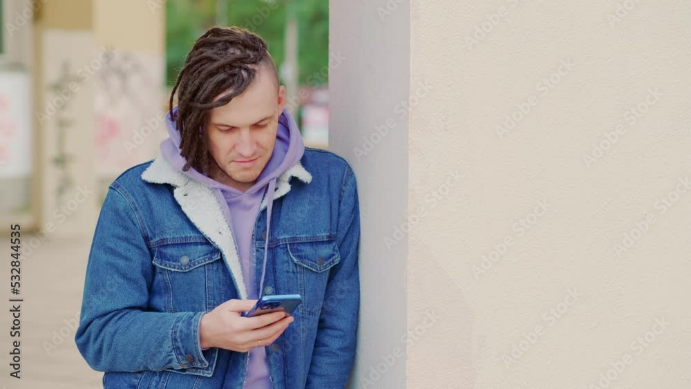 Portrait of young handsome man with dreadlocks browsing mobile phone, standing, leaning against wall on street. Portrait of young handsome man with dreadlocks browsing mobile phone, standing, leaning against wall on street.