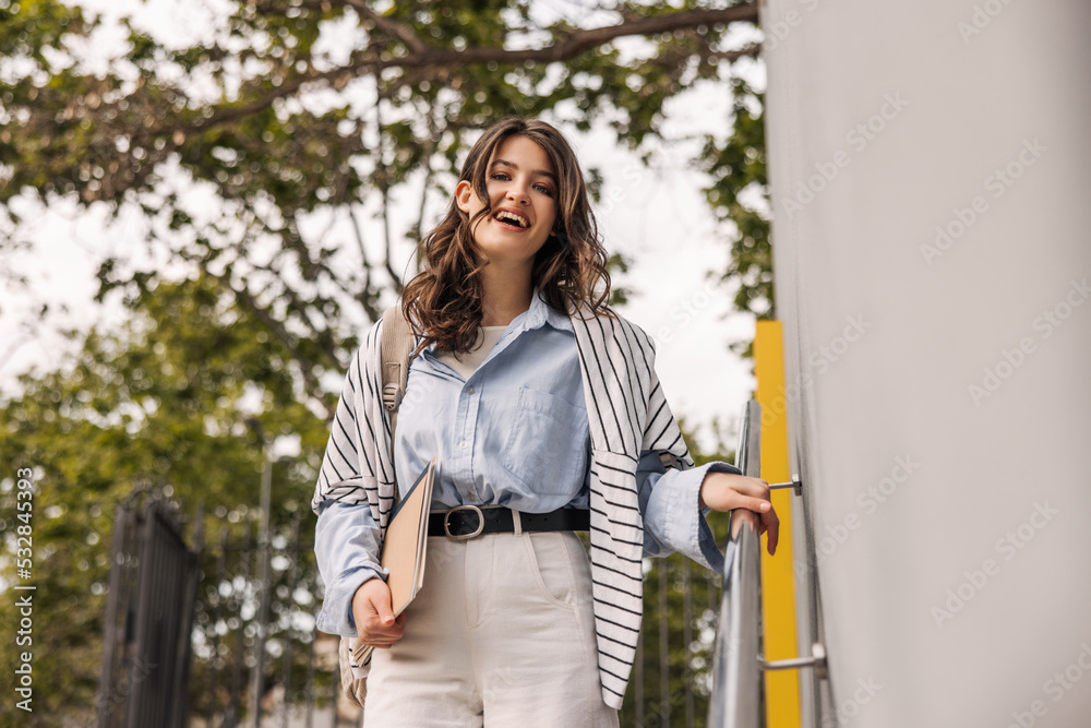 Happy young caucasian woman in good mood looks at camera going down stairs. Brunette with wavy hair wears casual clothes. Lifestyle concept Happy young caucasian woman in good mood looks at camera going down stairs. Brunette with wavy hair wears casual clothes. Lifestyle concept