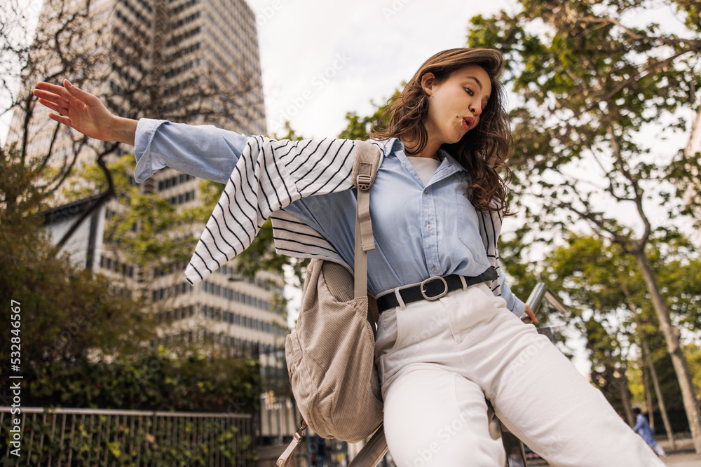Funny young caucasian woman with backpack having fun outdoors during day. Brunette wears shirt, sweatshirt and trousers to street. City life concept