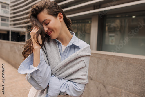 Cute young caucasian woman closing eyes embarrassedly covers face with hair standing outdoors. Brunette wears classic style of clothing. Leisure, relaxation and lifestyle concept. photo
