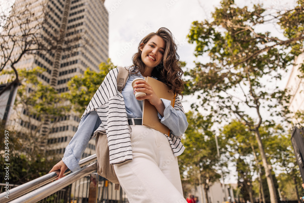 Bottom view happy young caucasian woman looking at camera, holding documents and coffee standing outdoors. Brunette with wavy hair wears casual clothes. Lifestyle concept