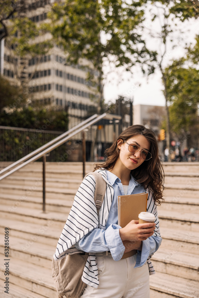Nice european young lady holding coffee, notebook going to college in morning. Brunette with wavy hair wears sunglasses, backpack, shirt and trousers. Concept of learning