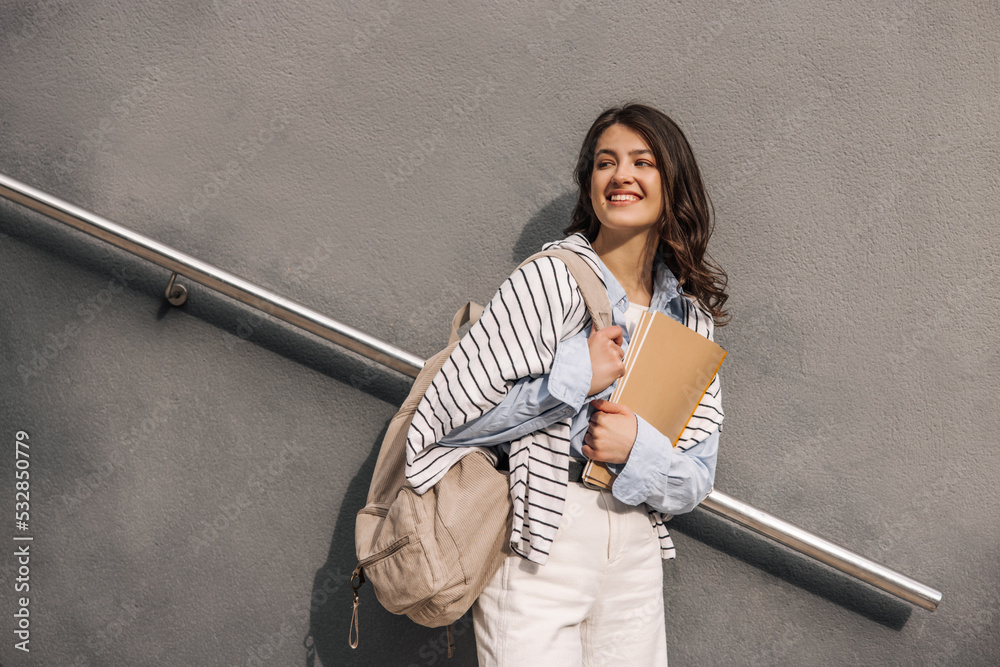 Stylish young caucasian female student with notebook and backpack looking away on grey background. Brunette woman with wavy hair wears classic clothes. Lifestyle concept Stylish young caucasian female student with notebook and backpack looking away on grey background. Brunette woman with wavy hair wears classic clothes. Lifestyle concept