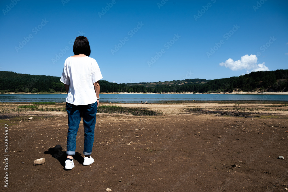 Caucasian woman from behind and standing on the shore of a lake, enjoying the scenery on a summer's day.
