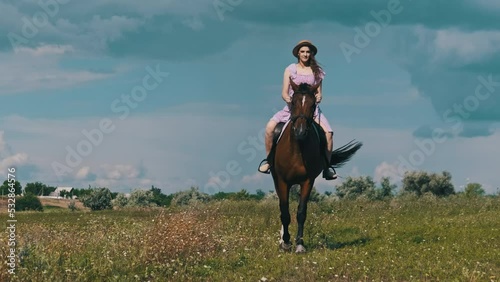 Young woman in a summer dress and straw hat riding a horse in a rural field in the countryside, slow motion. Horseback riding at equestrian yard. Attractive horsewoman is on a saddle riding. Lifestyle photo