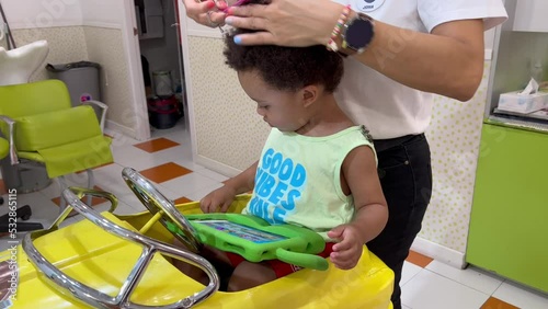 Exotic two year old african europoean  child in the hairdresser for his first hair cut, seated in a yellow toy car. photo