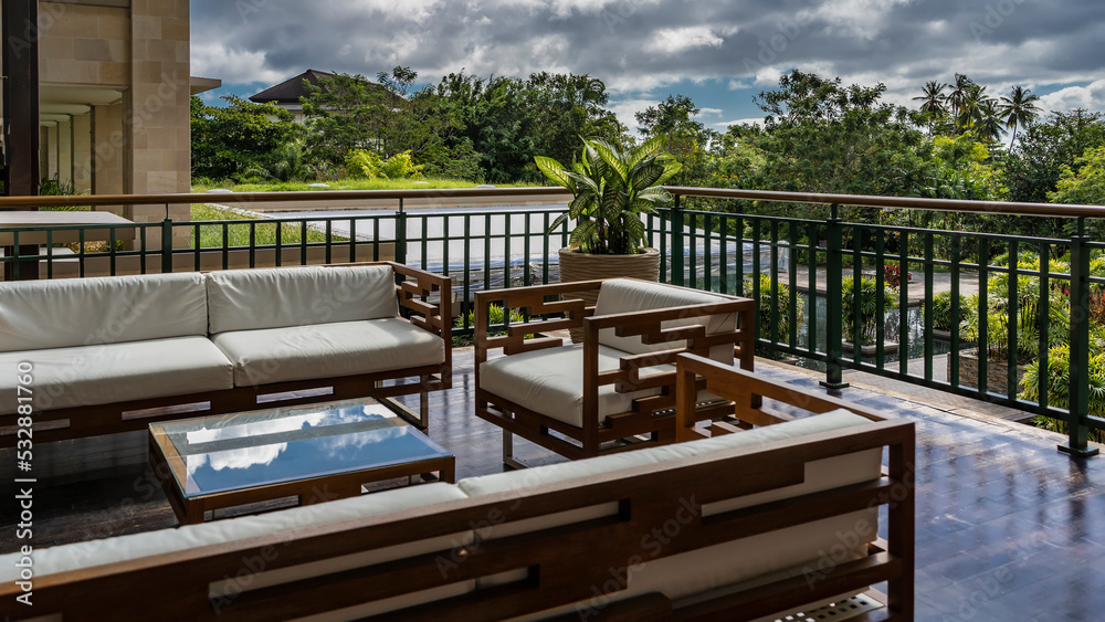 A seating area on the terrace. Wooden chairs and sofas with soft cushions are fenced with metal railings. The reflection of the sky on the glass of the coffee table. Green vegetation in the garden. 