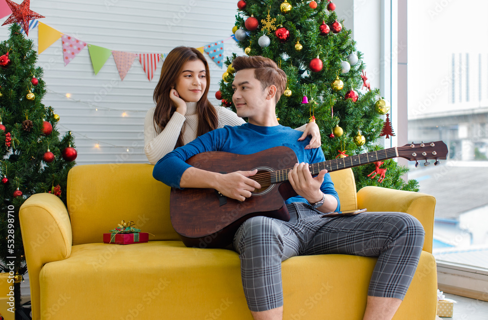 Asian happy cheerful romantic lover couple girlfriend smiling hugging cuddling boyfriend sitting on cozy sofa playing guitar sing song celebrating Christmas eve in full decorated home living room
