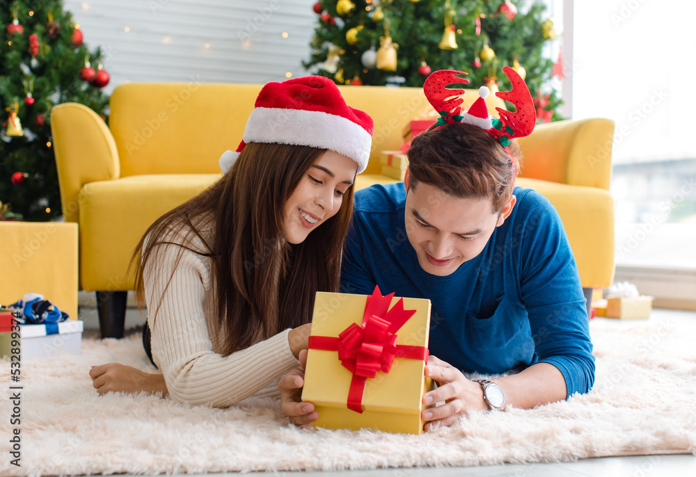 Asian cheerful boyfriend wears deer antler headband and girlfriend in sweater and red Santa Claus hat laying down on floor looking each other eyes in decorated living room celebrating Christmas eve Asian cheerful boyfriend wears deer antler headband and girlfriend in sweater and red Santa Claus hat laying down on floor looking each other eyes in decorated living room celebrating Christmas eve