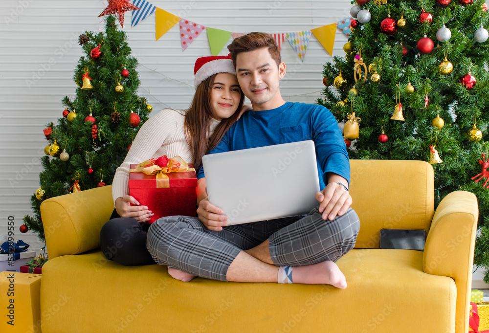 Asian happy boyfriend and girlfriend in sweater and red Santa Claus hat sitting on sofa together waving hand greeting family via laptop computer video call celebrating Christmas eve in decorated home Asian happy boyfriend and girlfriend in sweater and red Santa Claus hat sitting on sofa together waving hand greeting family via laptop computer video call celebrating Christmas eve in decorated home