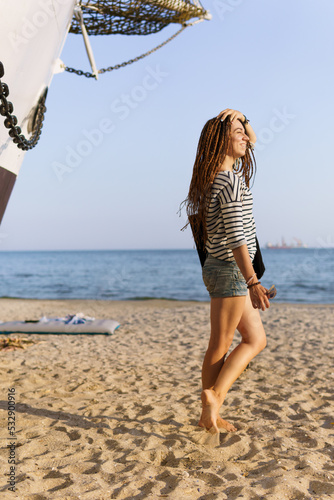 a girl with a dreadlocked hairstyle poses on the beach near a ship in summer, the sea and sand on a bright sunny day, dressed in a T-shirt and denim shorts