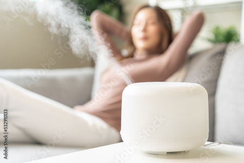 Modern air humidifier during relax or rest, happy blurred asian young woman, girl enjoying aromatherapy steam scent from essential oil diffuser comfortable in living conditions room,apartment at home