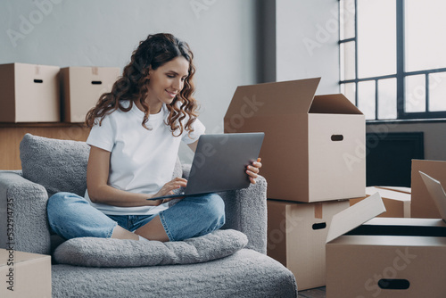 Smiling hispanic woman using laptop choosing moving service for moving in new home on relocation day photo
