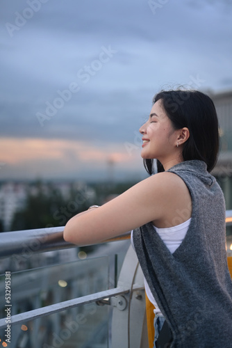 Relaxed Asian woman standing rooftop terrace in metropolis and looking cityscape at beautiful sunset