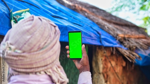 Old Indian man taking selfie and showing a green screen cell phone with rural village life concept background.