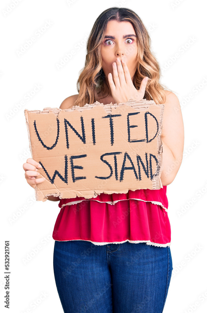 Young caucasian woman holding united we stand banner covering mouth with hand, shocked and afraid for mistake. surprised expression Young caucasian woman holding united we stand banner covering mouth with hand, shocked and afraid for mistake. surprised expression