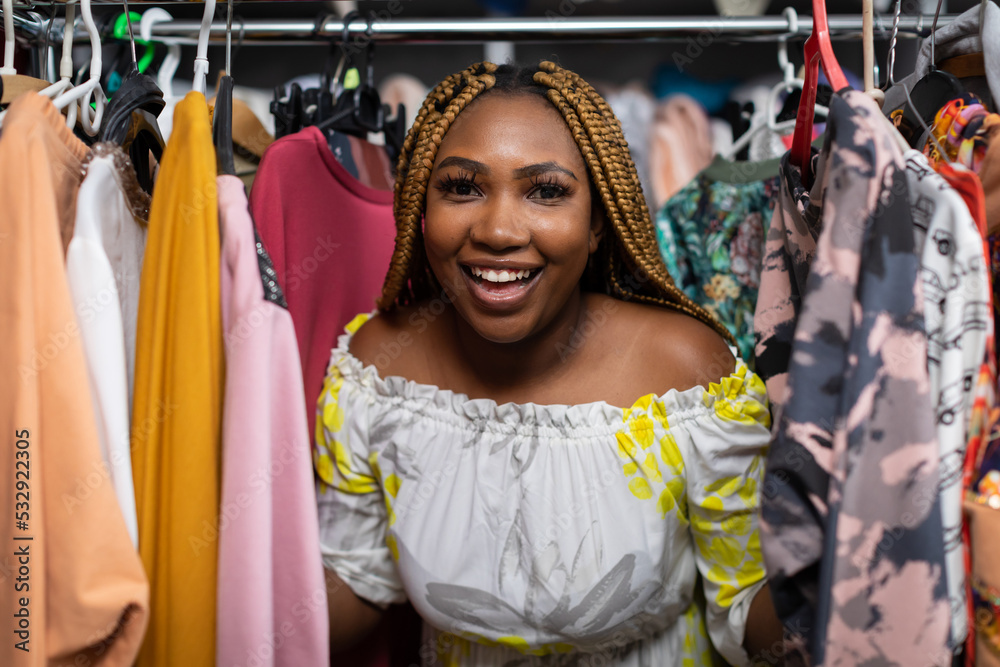 A smiling dark-skinned woman leans out from behind a rack full of clothing.