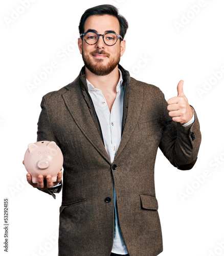Young hispanic man holding piggy bank smiling happy and positive, thumb up doing excellent and approval sign photo