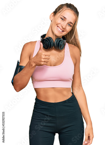 Fototapeta Naklejka Na Ścianę i Meble -  Young blonde woman wearing gym clothes and using headphones doing happy thumbs up gesture with hand. approving expression looking at the camera showing success.