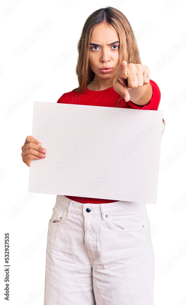 Young beautiful blonde woman holding blank empty banner pointing with finger to the camera and to you, confident gesture looking serious