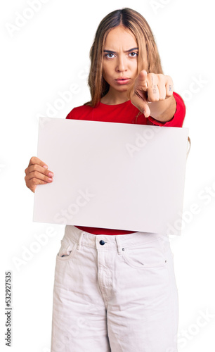 Young beautiful blonde woman holding blank empty banner pointing with finger to the camera and to you, confident gesture looking serious photo