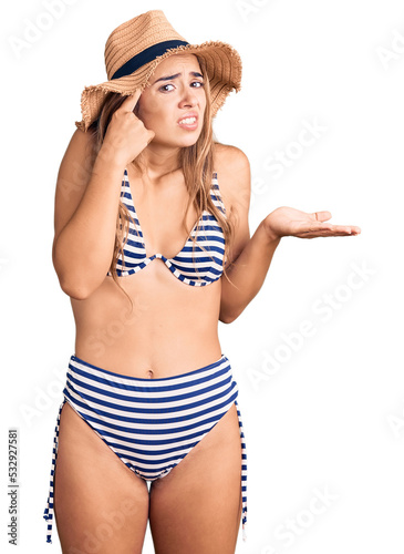 Young beautiful blonde woman wearing bikini and hat confused and annoyed with open palm showing copy space and pointing finger to forehead. think about it.