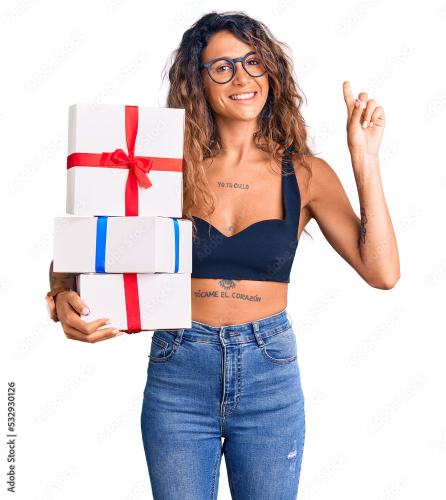 Young hispanic woman with tattoo holding gifts surprised with an idea or question pointing finger with happy face, number one Young hispanic woman with tattoo holding gifts surprised with an idea or question pointing finger with happy face, number one