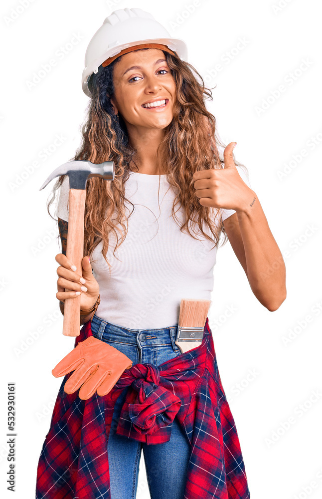 Young hispanic woman with tattoo wearing hardhat and builder clothes holding hammer smiling happy and positive, thumb up doing excellent and approval sign Young hispanic woman with tattoo wearing hardhat and builder clothes holding hammer smiling happy and positive, thumb up doing excellent and approval sign