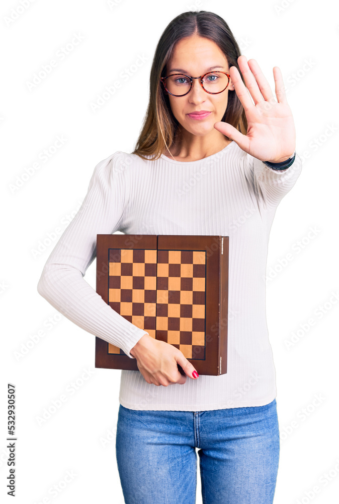 Beautiful brunette young woman holding chess with open hand doing stop sign with serious and confident expression, defense gesture Beautiful brunette young woman holding chess with open hand doing stop sign with serious and confident expression, defense gesture