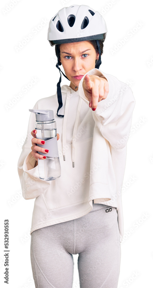 Beautiful brunette young woman wearing bike helmet and holding water bottle pointing with finger to the camera and to you, confident gesture looking serious Beautiful brunette young woman wearing bike helmet and holding water bottle pointing with finger to the camera and to you, confident gesture looking serious