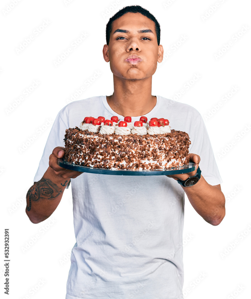 Young hispanic man celebrating birthday holding big chocolate cake puffing cheeks with funny face. mouth inflated with air, catching air.