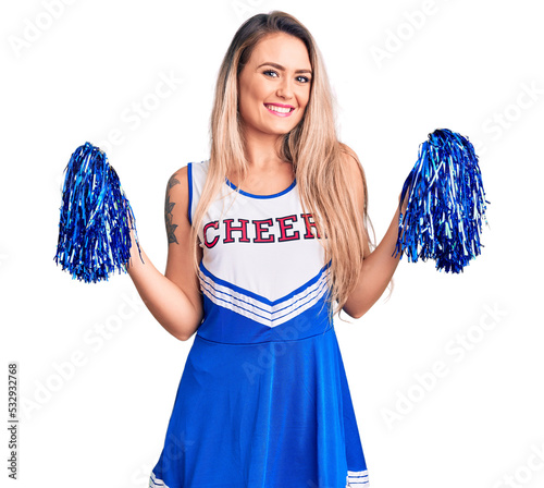 Young beautiful blonde woman wearing cheerleader uniform holding pompom looking positive and happy standing and smiling with a confident smile showing teeth