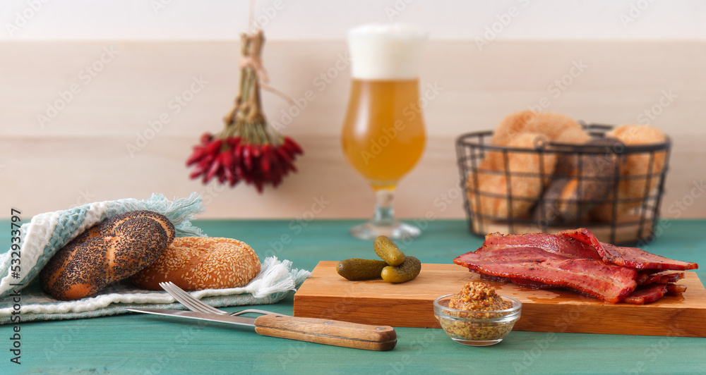 Meat snacks, grilled bacon slices and cornichons served on a wooden cutting board, coarse mustard, bread and a glass of unfiltered beer.
