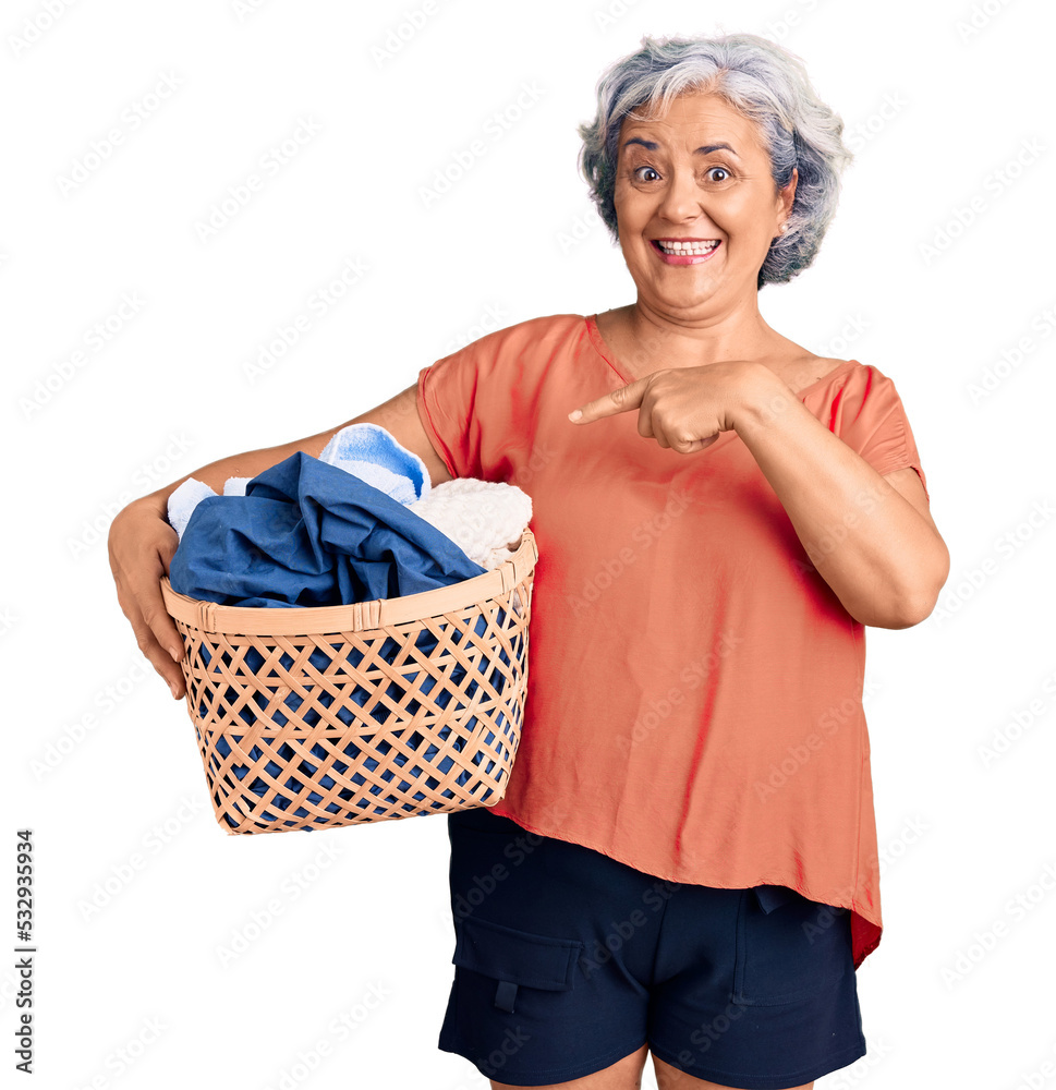 Senior woman with gray hair holding laundry basket smiling happy pointing with hand and finger Senior woman with gray hair holding laundry basket smiling happy pointing with hand and finger