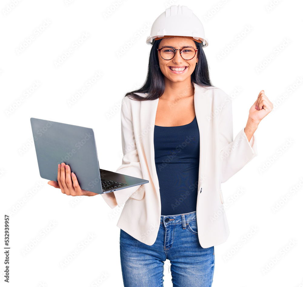 Young beautiful latin girl wearing architect hardhat holding laptop screaming proud, celebrating victory and success very excited with raised arms Young beautiful latin girl wearing architect hardhat holding laptop screaming proud, celebrating victory and success very excited with raised arms