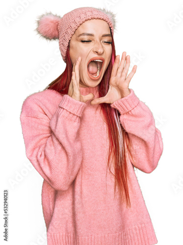 Young caucasian woman wearing casual clothes and wool cap shouting angry out loud with hands over mouth
