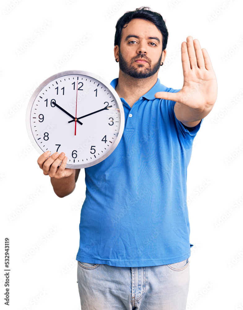 Young hispanic man holding big clock with open hand doing stop sign with serious and confident expression, defense gesture Young hispanic man holding big clock with open hand doing stop sign with serious and confident expression, defense gesture