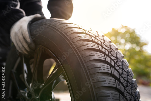 Close-up of hands with gloves rolling a new winter tyre  in bright sunlight ready for tyre change. Driving safety, road safety and car service concept.