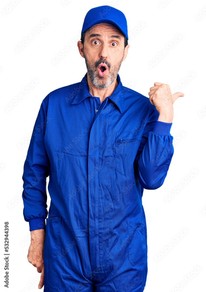 Middle age handsome man wearing mechanic uniform surprised pointing with hand finger to the side, open mouth amazed expression.