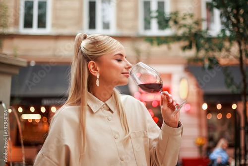 Charming young woman in beige coat on evening street of an old European city tastes and enjoys red wine © okostia