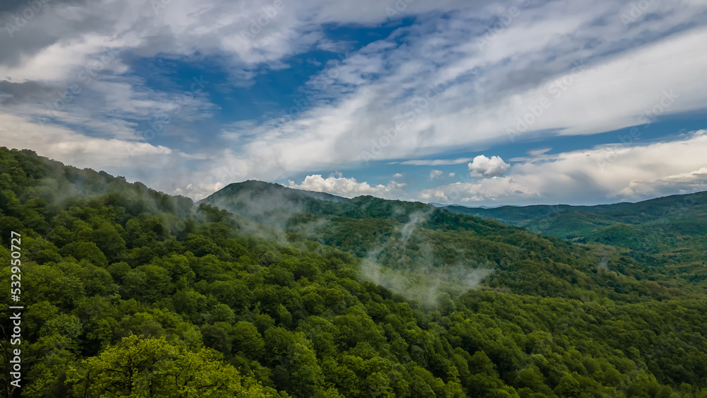 Beautiful nature in the mountains. Fog over the forests. Blue sky with clouds. Green trees on the slopes of the mountains. Mystical fog after the rain.