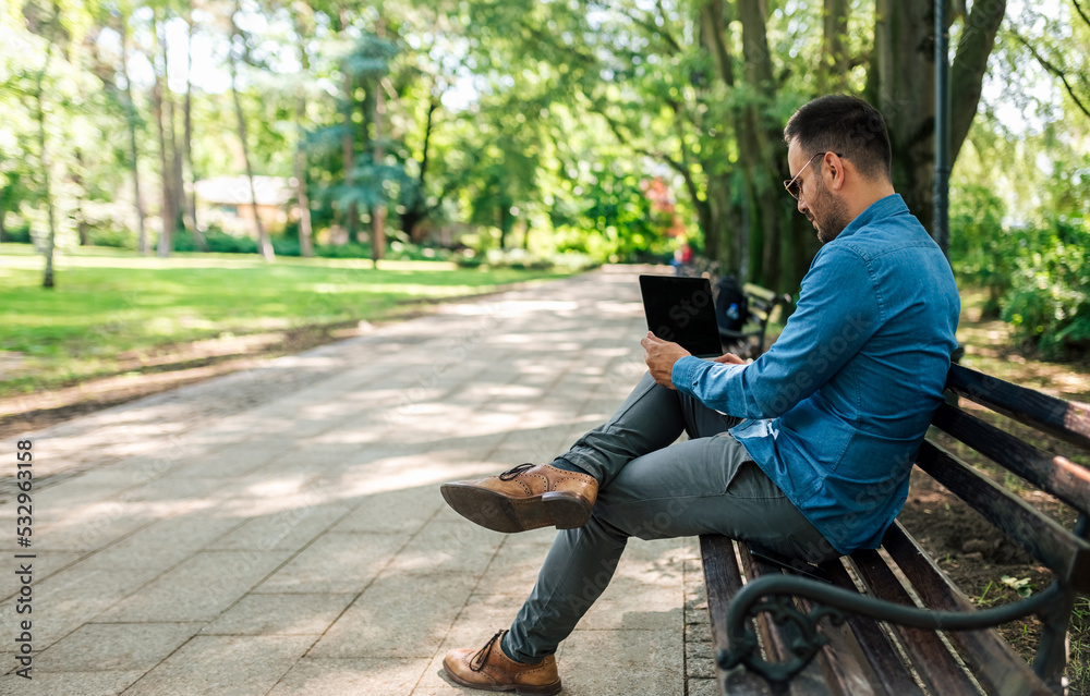 Obraz premium Serious young adult businessman, sitting in the park, turning on his laptop.