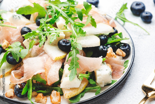 Tasty arugula salad with sweet pears, blueberries, roquefort cheese, smoked pork ham and crunchy walnuts. White kitchen table background, top view, space for text