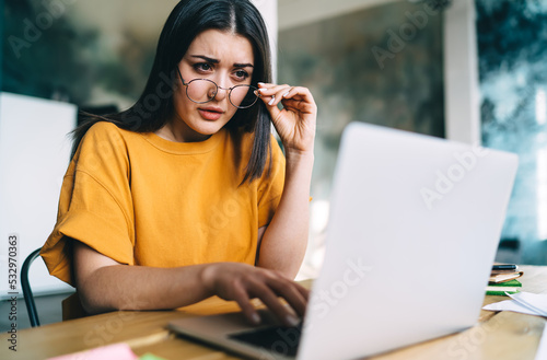 Unsure young woman in glasses looking at screen of netbook