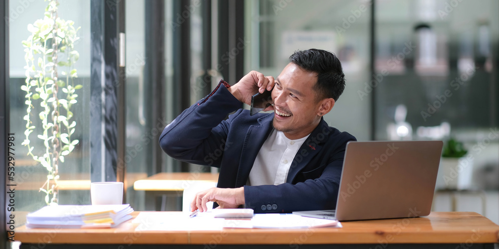 Asian businessman sitting on the phone with a customer with a laptop and document at his office desk. Asian businessman sitting on the phone with a customer with a laptop and document at his office desk.
