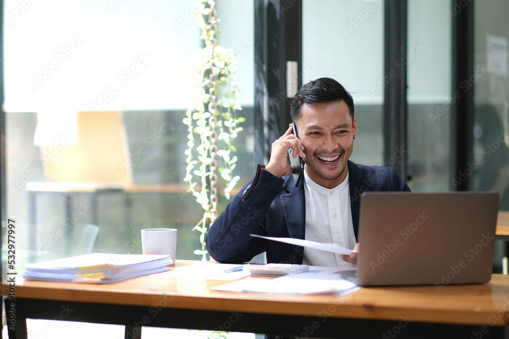 Asian businessman sitting on the phone with a customer with a laptop and document at his office desk. Asian businessman sitting on the phone with a customer with a laptop and document at his office desk.